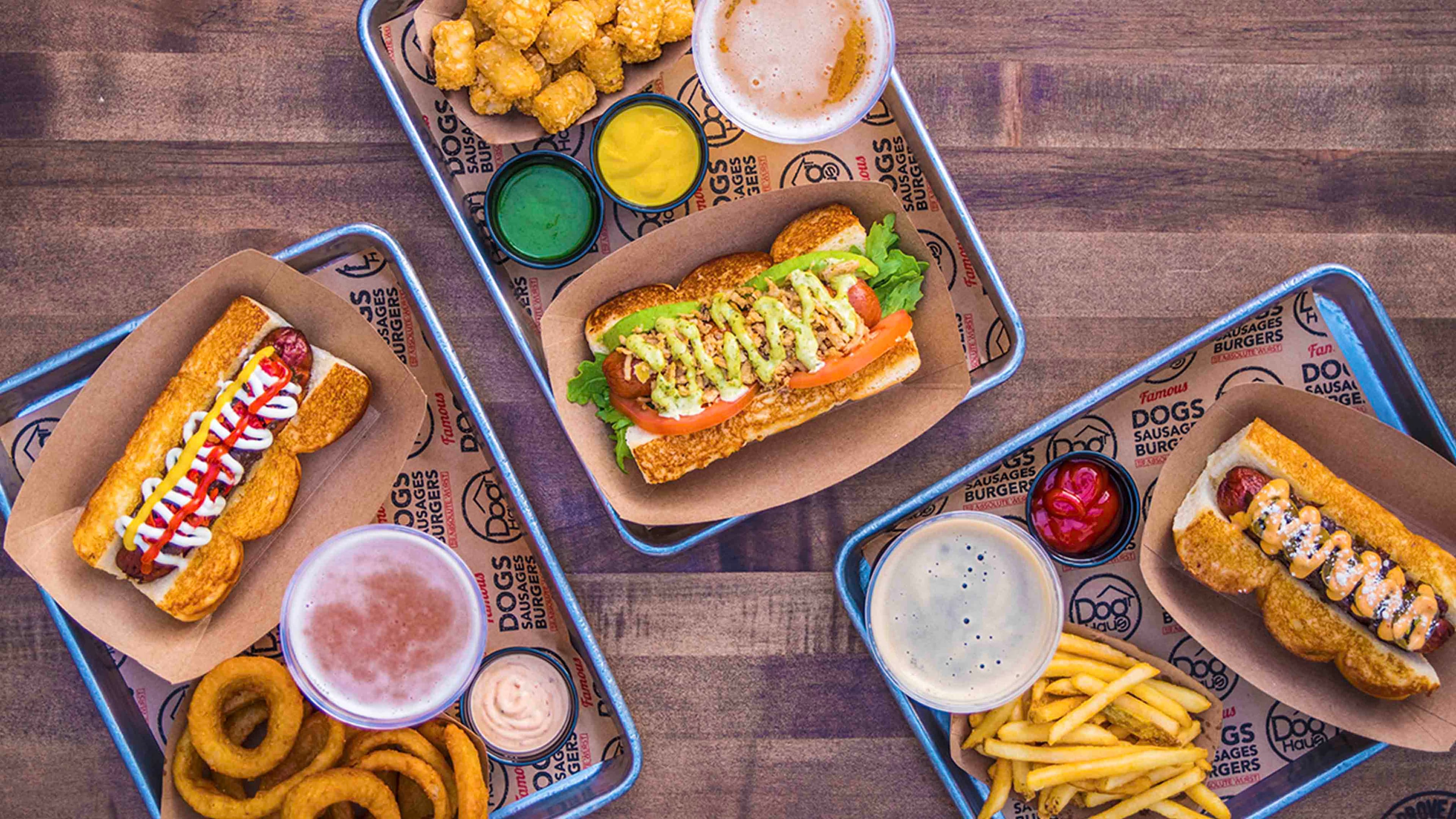 An overhead view of multiple Dog Haus trays filled with gourmet hot dogs, tater tots, onion rings, French fries, and dipping sauces, served on branded paper with beers on a wooden table.